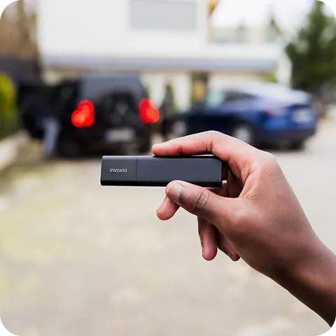 Un Classic GPS Tracker tenu dans une main, avec un arrière-plan flou montrant deux voitures dans une cour